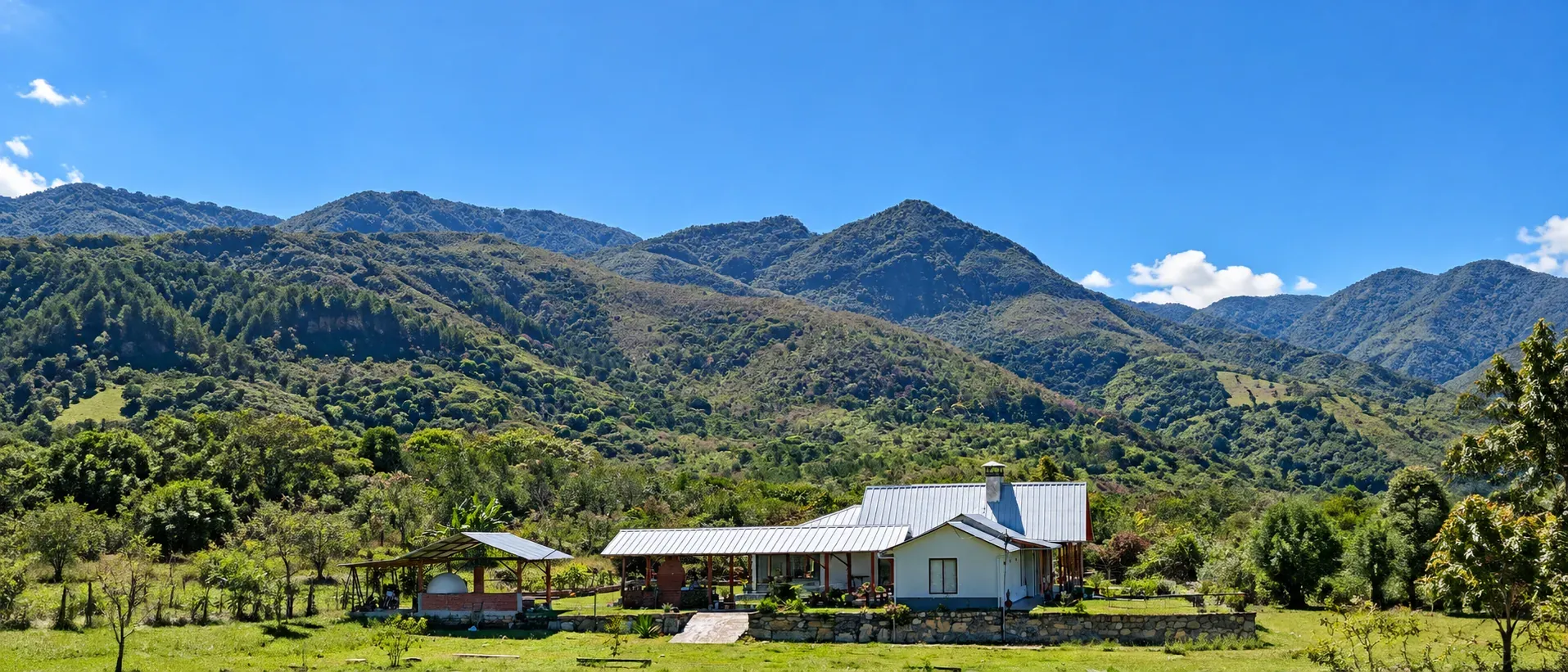 Una casa de techo metálico entre pastos verdes, con montañas boscosas y cielo azul detrás.