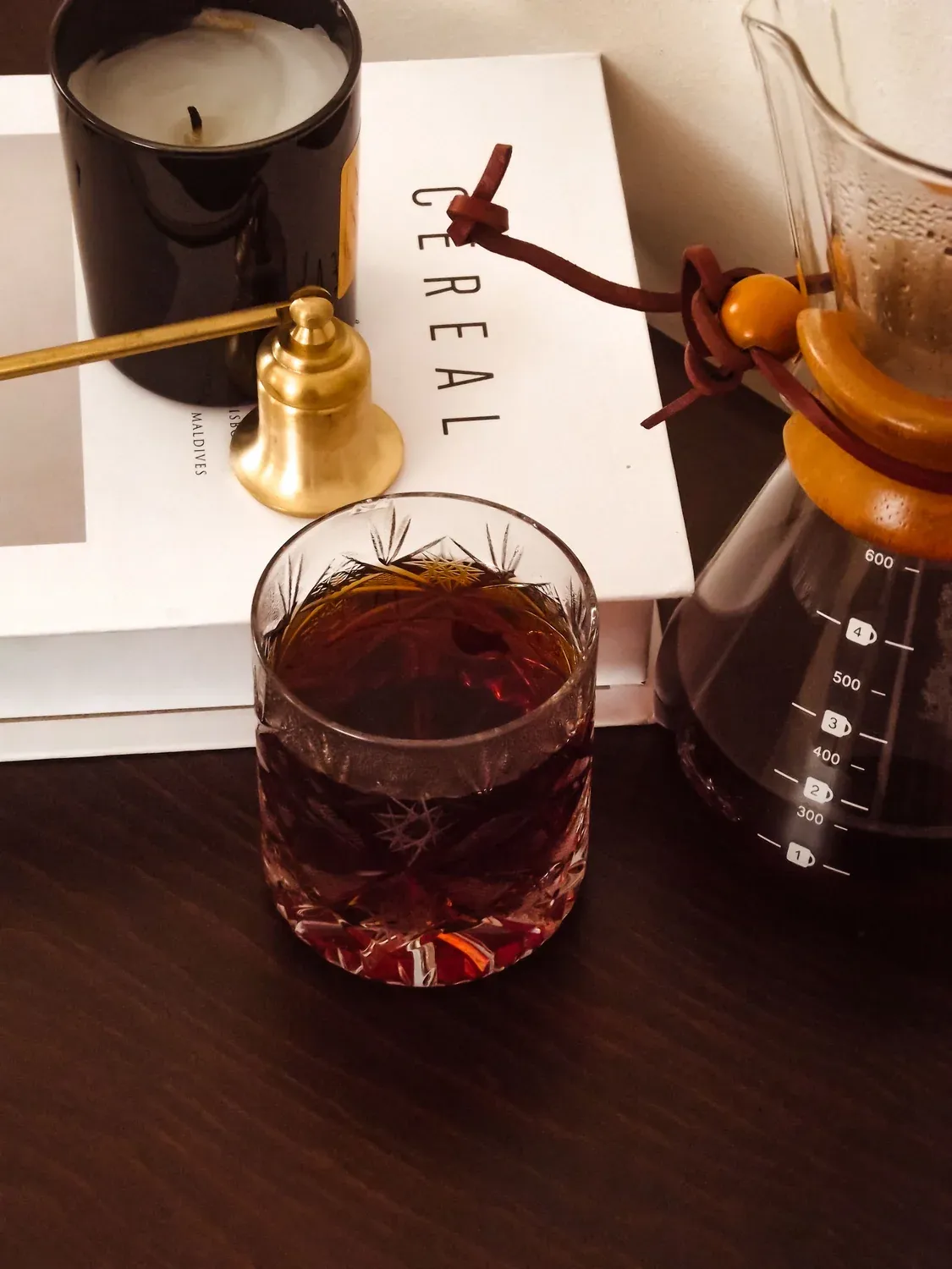 Un vaso de café oscuro junto a una jarra Chemex, una vela y un libro.