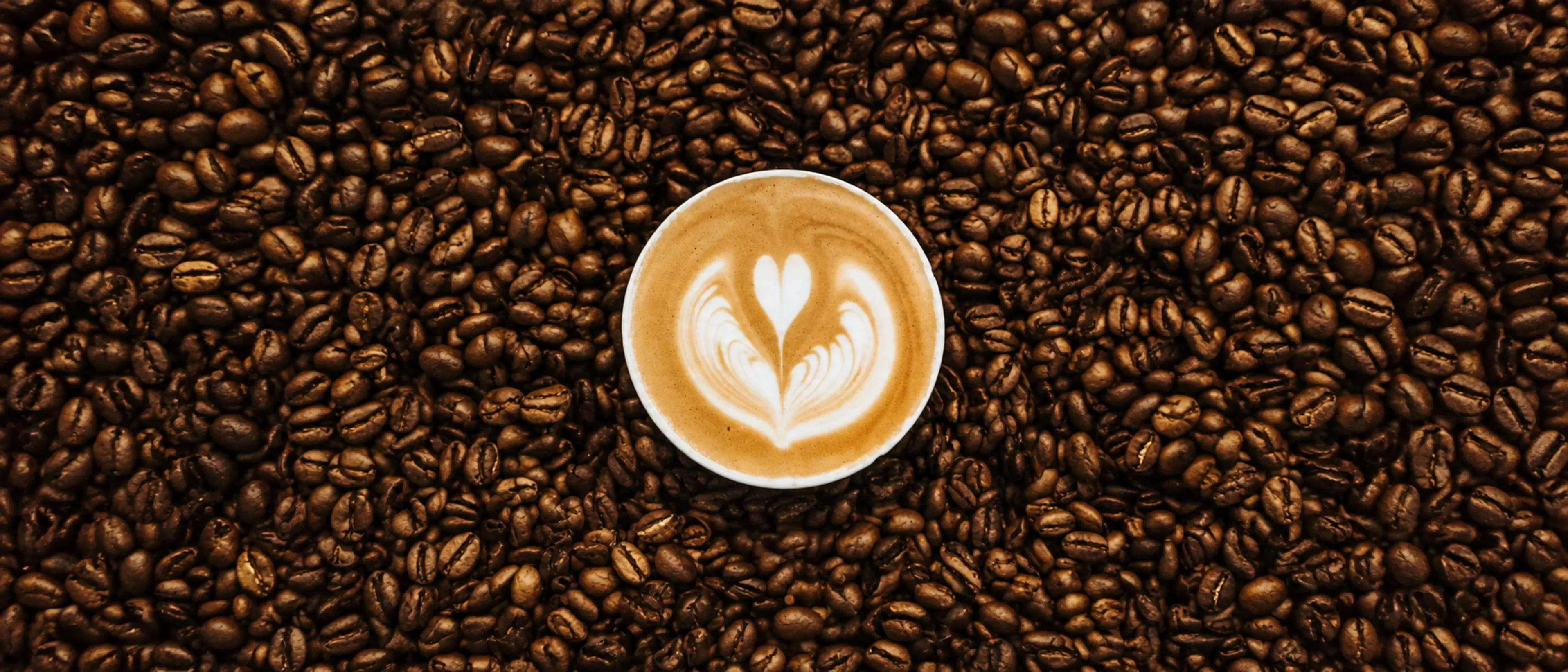 Vista desde arriba de una taza de café con arte latte en forma de corazón, rodeada de granos de café tostado.