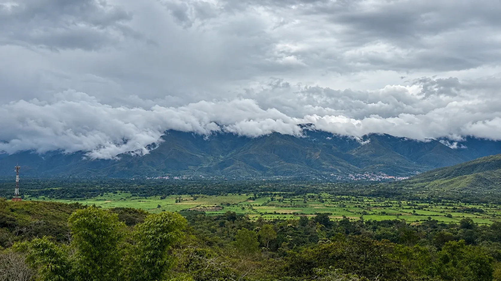 Valle de campos verdes bajo montañas cubiertas por nubes, con un poblado a lo lejos.